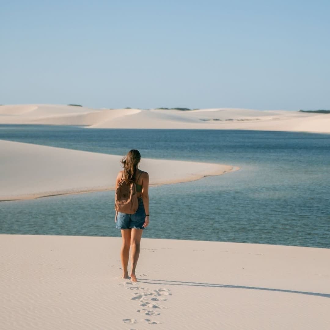 Passos na areia fina, lagoa à frente, tempo de sobra. Uma mulher com mochila caminha descalça sobre dunas de areia branca em direção a uma lagoa azul cristalina no roteiro completo dos Lençóis Maranhenses, com suas pegadas visíveis na areia atrás dela