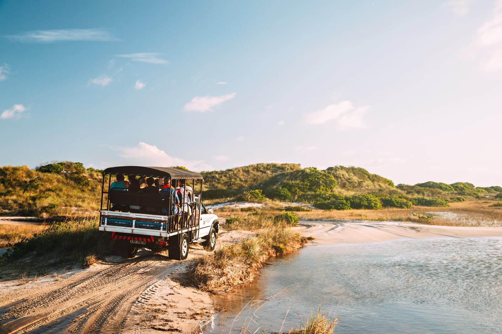 Le chemin compte autant que la destination, surtout quand le lagon est à portée de main. Un 4x4 à toit ouvert transporte un groupe de visiteurs sur une piste sablonneuse longeant une lagune peu profonde, avec des dunes végétalisées en arrière-plan sous un ciel bleu à Lençóis Maranhenses près de Santo Amaro