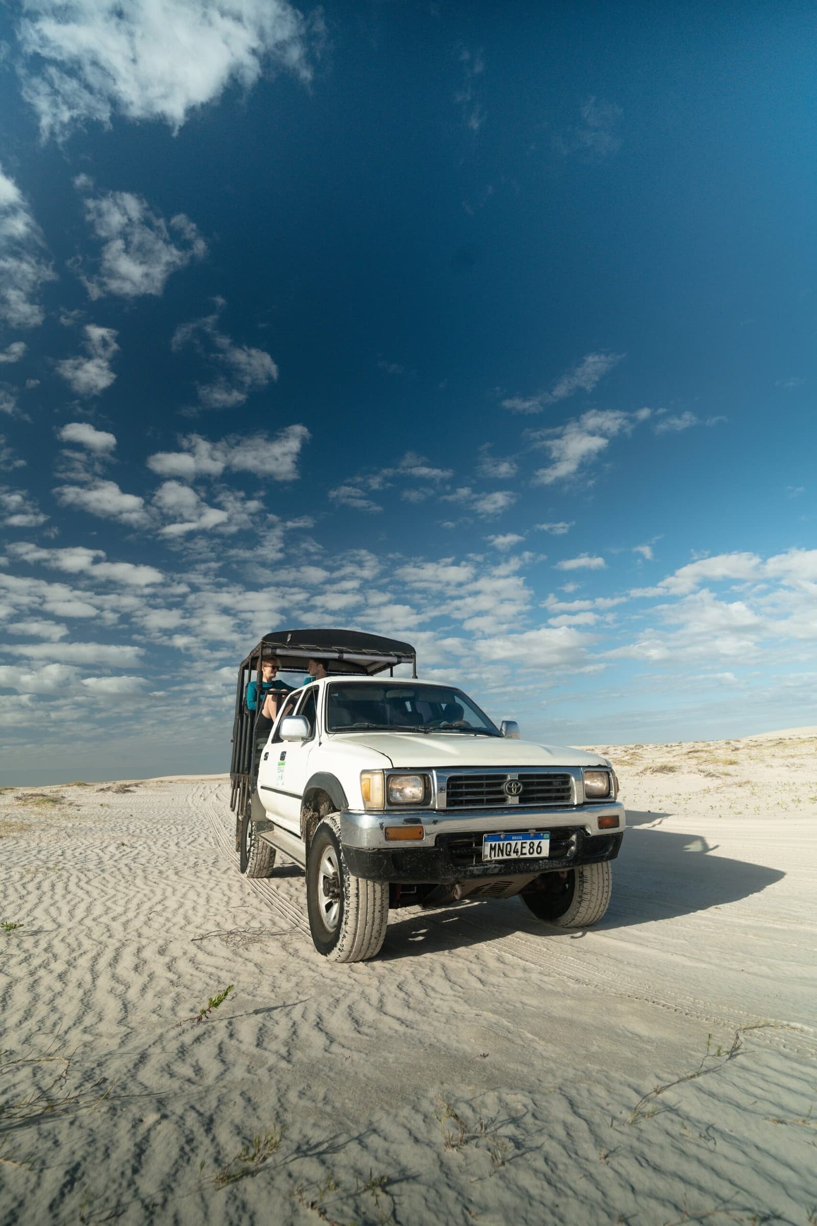 Assim é chegar aos Lençóis Maranhenses: de picape, janela aberta e sem pressa. Uma Toyota 4x4 branca com caçamba aberta e passageiros cruza uma vasta planície de dunas brancas sob um céu azul intenso durante o trek pelos Lençóis Maranhenses.