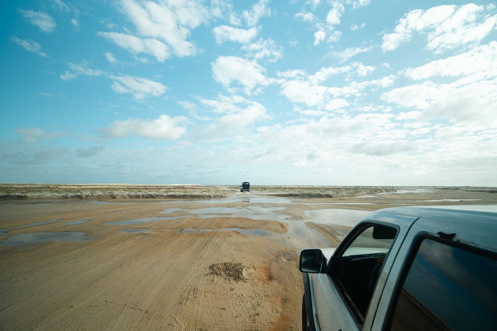 Por aqui, a estrada é o que a maré resolveu deixar. Dois veículos 4x4 cruzando uma ampla planície arenosa e úmida sob um céu azul parcialmente nublado durante o trek pelos Lençóis Maranhenses saindo de Atins, com pequenas poças d'água refletindo a luz ao longo do terreno
