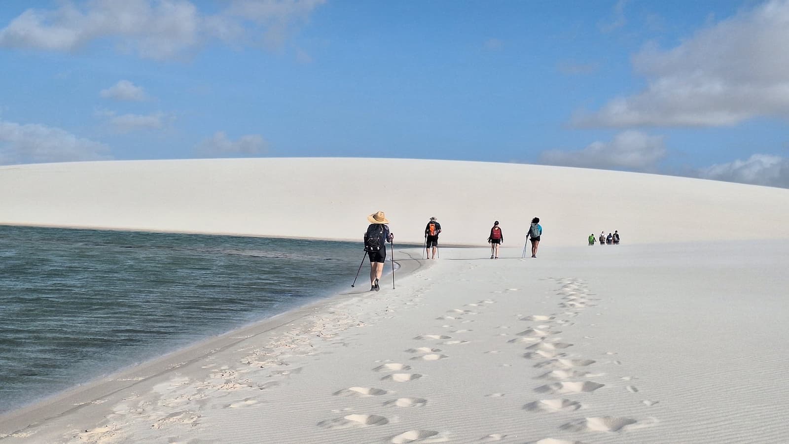 Areia de um lado, água do outro: o caminho se desenha sozinho. Um grupo de caminhantes com bastões e mochilas percorre uma estreita crista de areia branca à beira de uma lagoa de água doce verde-escura durante o trek pelos Lençóis Maranhenses saindo de Atins, com uma grande duna clara se curvando contra o céu azul à frente