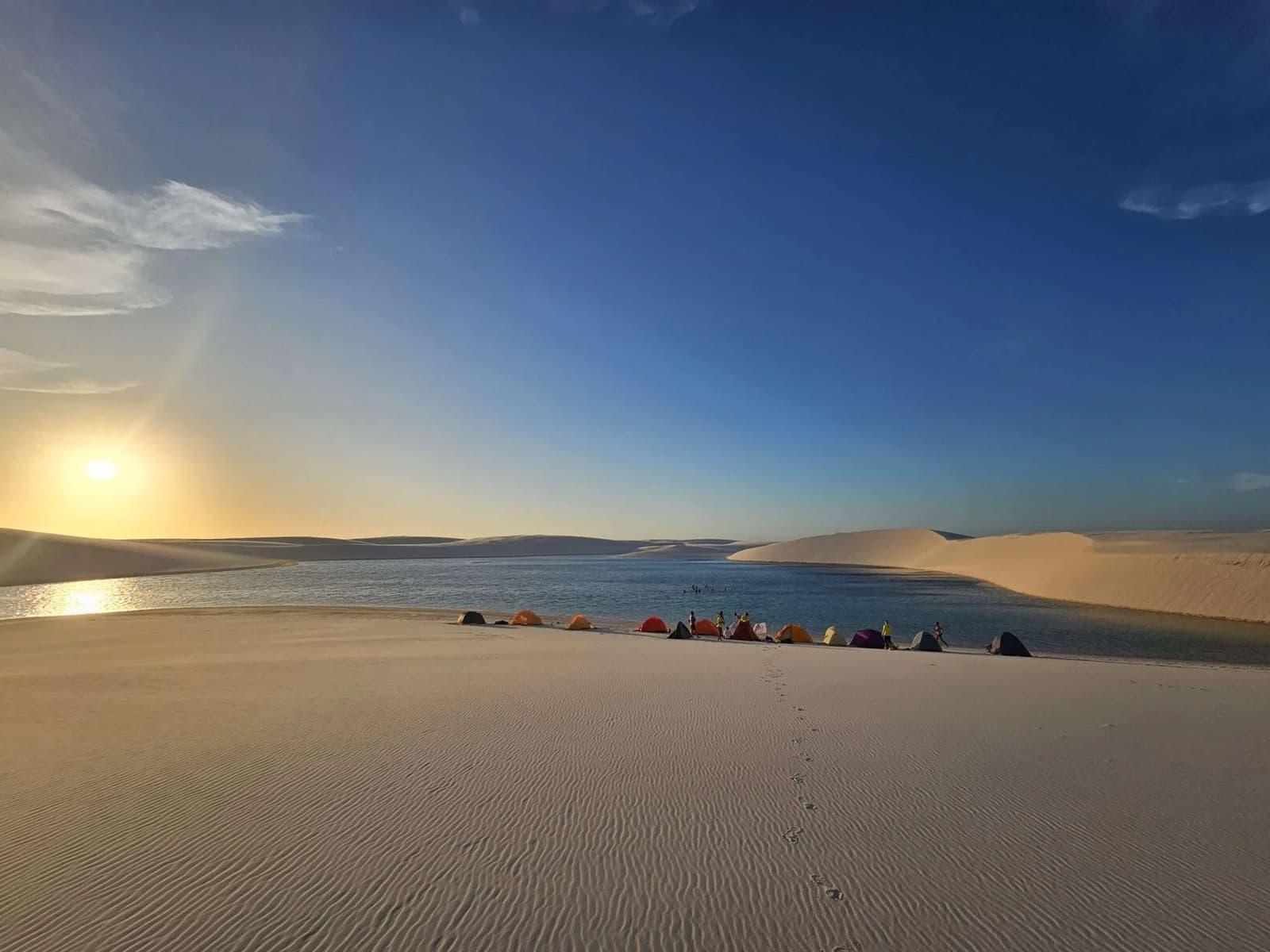 Acampar nos Lençóis é o tipo de experiência que muda o seu referencial de 'lindo'. Barracas coloridas enfileiradas à beira de uma lagoa de água doce ao pôr do sol durante o trek pelos Lençóis Maranhenses, com dunas de areia branca onduladas se estendendo ao fundo sob um céu azul intenso