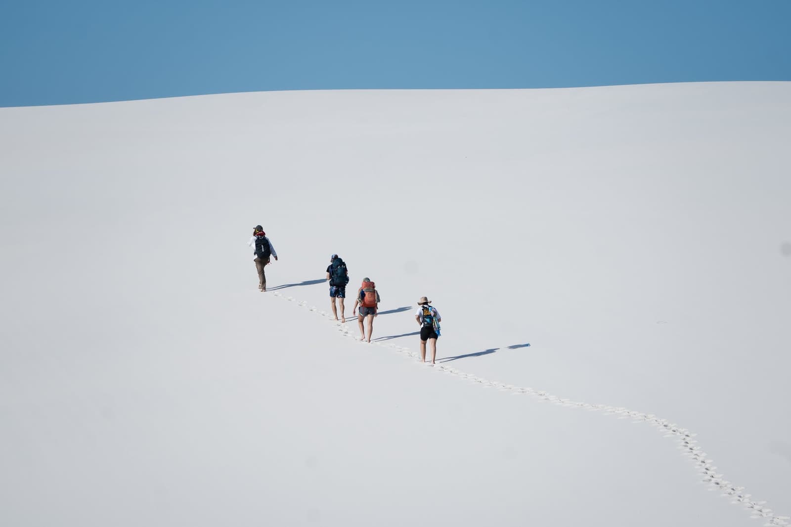 Cada pegada na areia branca é uma história que o vento vai apagar logo mais. Quatro caminhantes com mochilas sobem em fila uma enorme duna de areia branca durante o trek pelos Lençóis Maranhenses, deixando um rastro de pegadas na encosta vazia sob um céu azul límpido