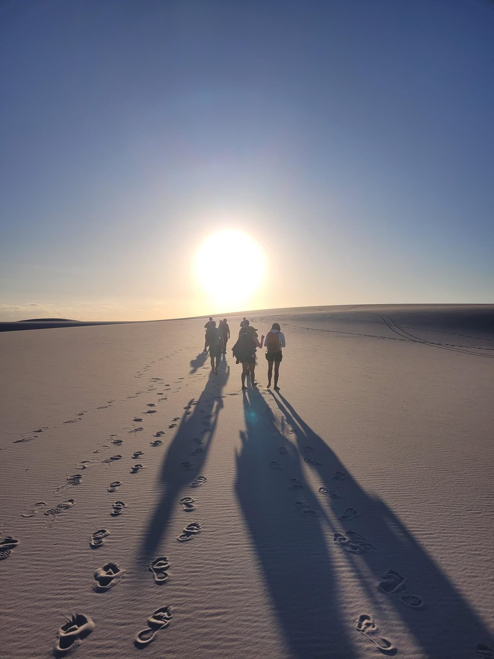 Areias brancas, sombras longas, e o dia se despedindo devagar. Um pequeno grupo de caminhantes avança em direção ao sol baixo nas dunas brancas durante o trek pelos Lençóis Maranhenses, com longas sombras e pegadas descalças desenhadas na areia