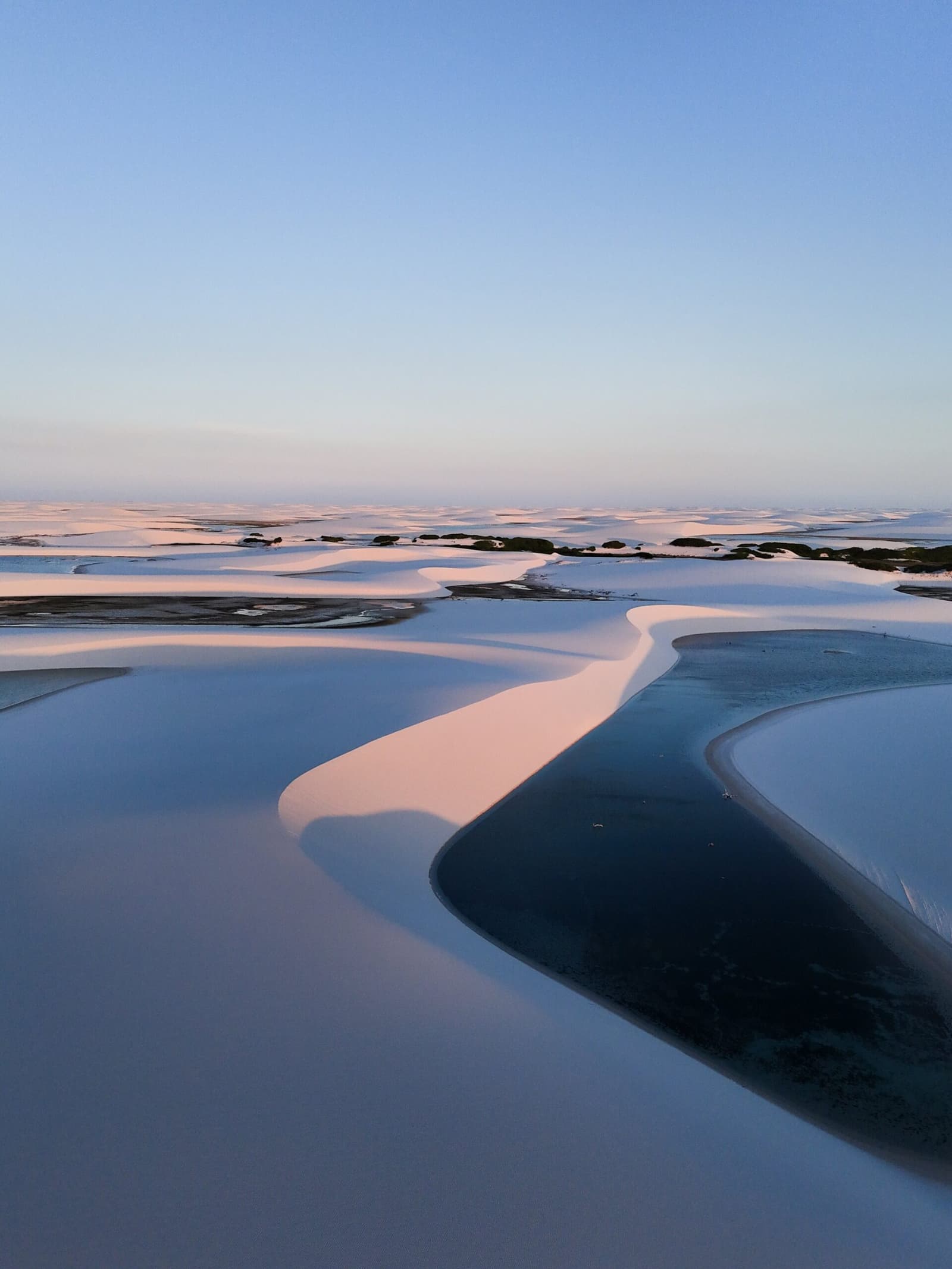 Le sable courbe, la lumière hésite, et on comprend enfin pourquoi on a fait le voyage. Vue aérienne de dunes de sable blanc et de lagunes bleu foncé au crépuscule sur le parcours de trekking des Lençóis Maranhenses, avec une lumière rose chaude sur les crêtes et un ciel pâle à l'horizon
