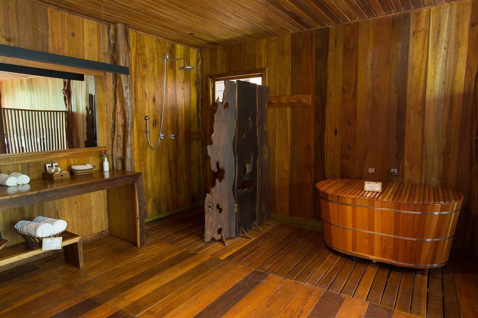 Le bois partout, l'eau chaude, et la forêt à portée de regard. Salle de bain tout en bois au Mirante do Gavião avec une baignoire tonneau, une douche à effet pluie, une cloison en bois brut et des serviettes blanches pliées sur un banc lamellé