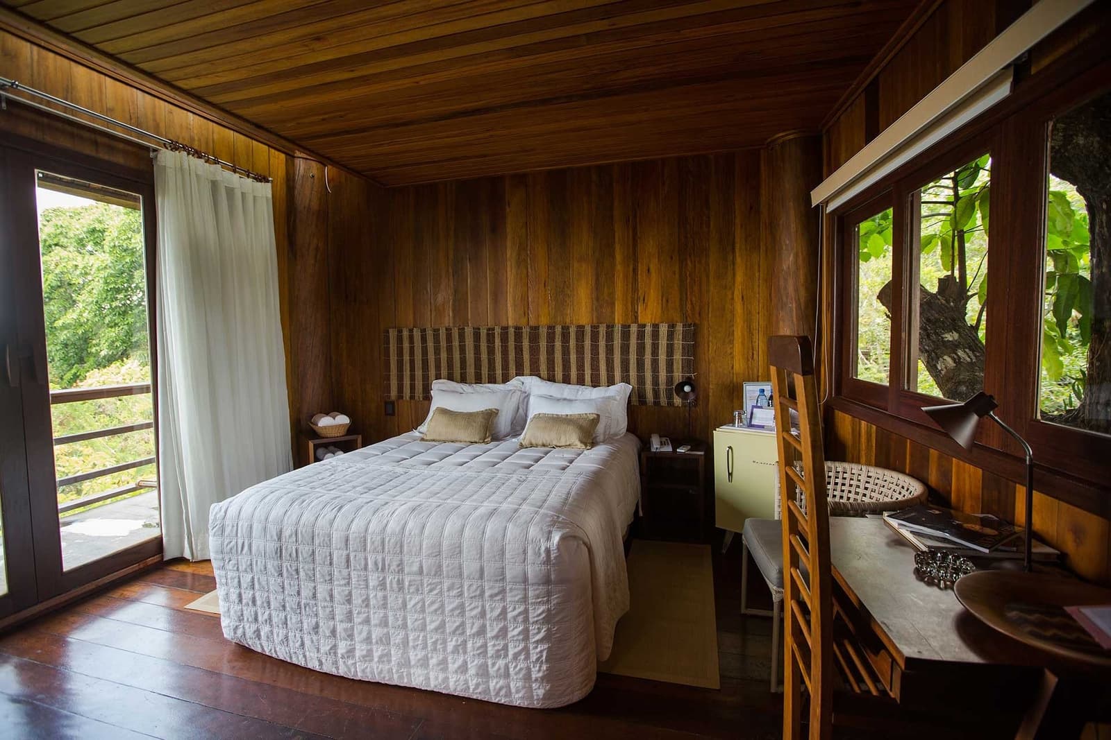 Le bois, le blanc, et la forêt qui s'invite par les fenêtres. Une chambre d'hôte du Mirante do Gavião avec des murs, un plafond et un plancher en bois chaleureux, un grand lit blanc piqué, une tête de lit tissée, un bureau en bois et des fenêtres donnant sur une végétation tropicale luxuriante