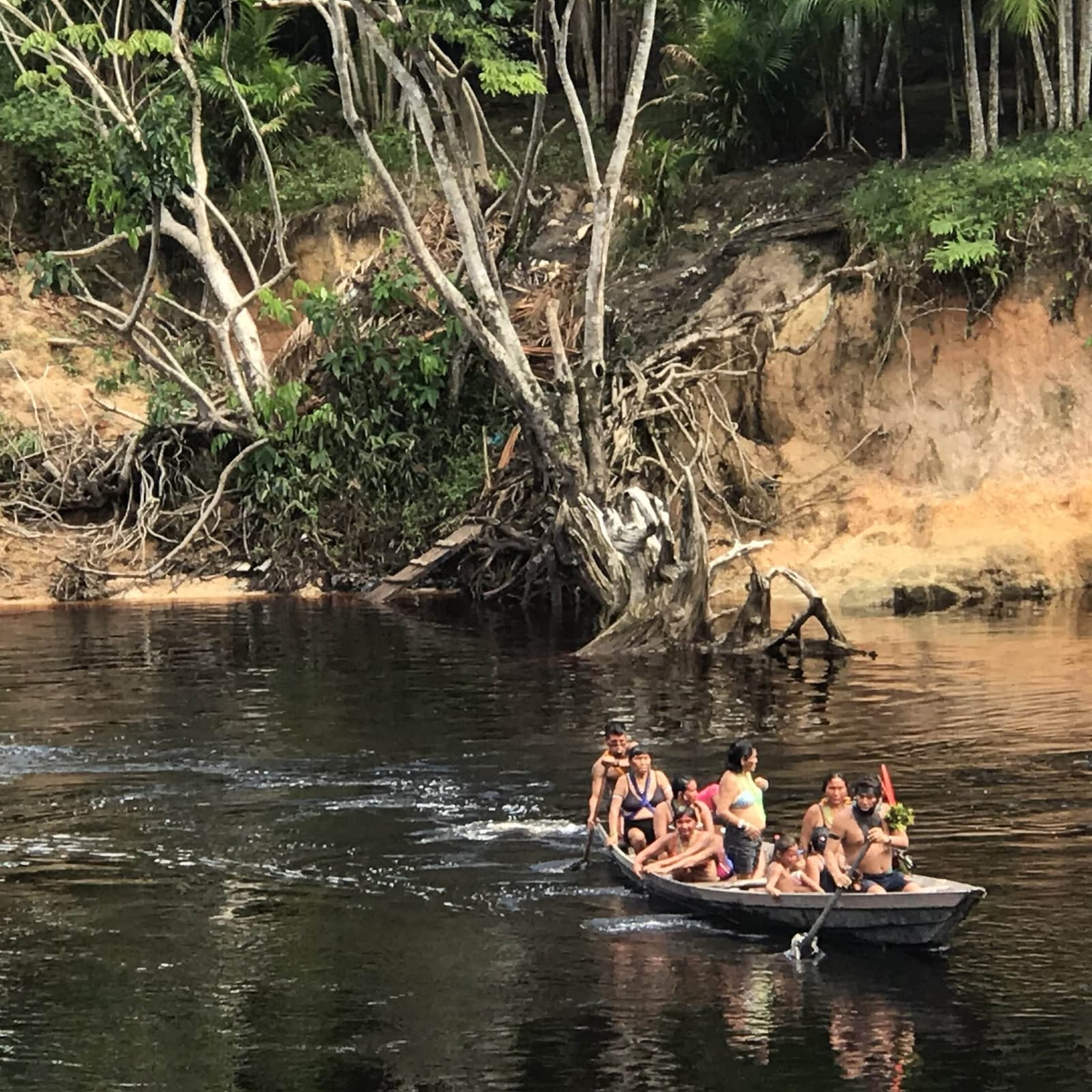 Op de rivier past er altijd nog iemand bij, dat is gewoon zo. Een vol houten motorbootje met een lokale familie vaart over een donker Amazone-rivierwater, met een geërodeerde kleiboever en dicht tropisch bos op de achtergrond.