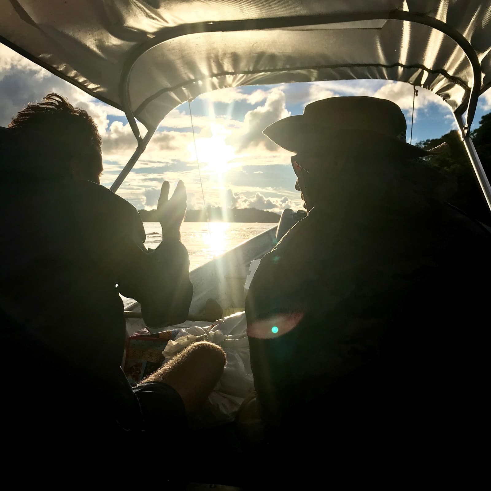 Vredesteken en tegenlicht, ergens diep in het noordwesten van Amazonia. Twee reizigers als silhouet in een overdekte motorboot tijdens de Pico da Neblina-expeditie, één met een vredesteken terwijl de zon door de wolken breekt boven een brede Amazone-rivier