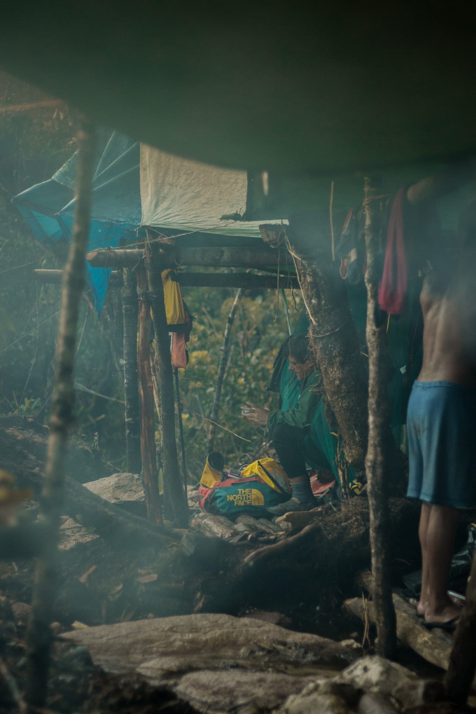 Rook, hoogte en een tas die betere dagen heeft gekend. Kamperen heeft hier niets glamoureus. Een trekker hurkt naast een gele North Face dry bag in een geïmproviseerde houten schuilplaats tijdens de Pico da Neblina Expeditie, rook trekt door de mistige lucht terwijl kleurrijke uitrusting aan bamboestokken hangt