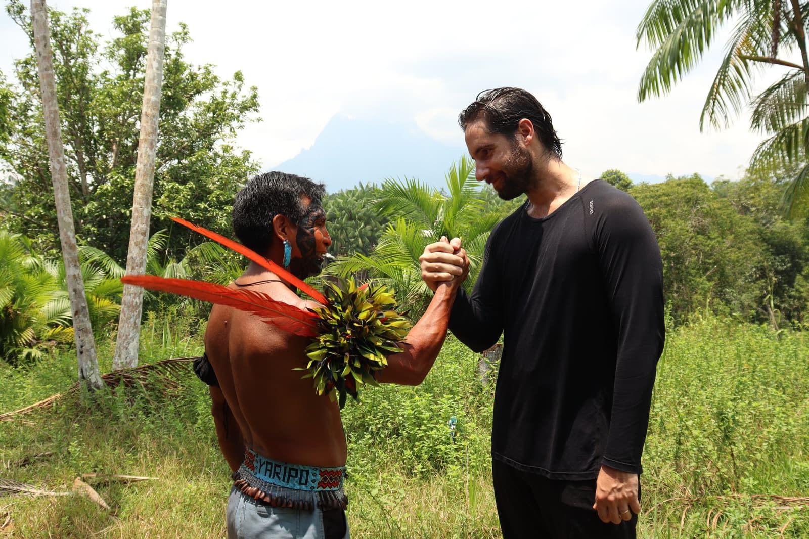 Een handdruk die geen uitleg nodig heeft. Een Yanomami-man met traditionele gezichtsbeschildering, rode veren en een bladversiering geeft een reiziger een hartelijke handdruk tijdens de Pico da Neblina Expeditie, met tropisch regenwoud en een bergtop op de achtergrond.