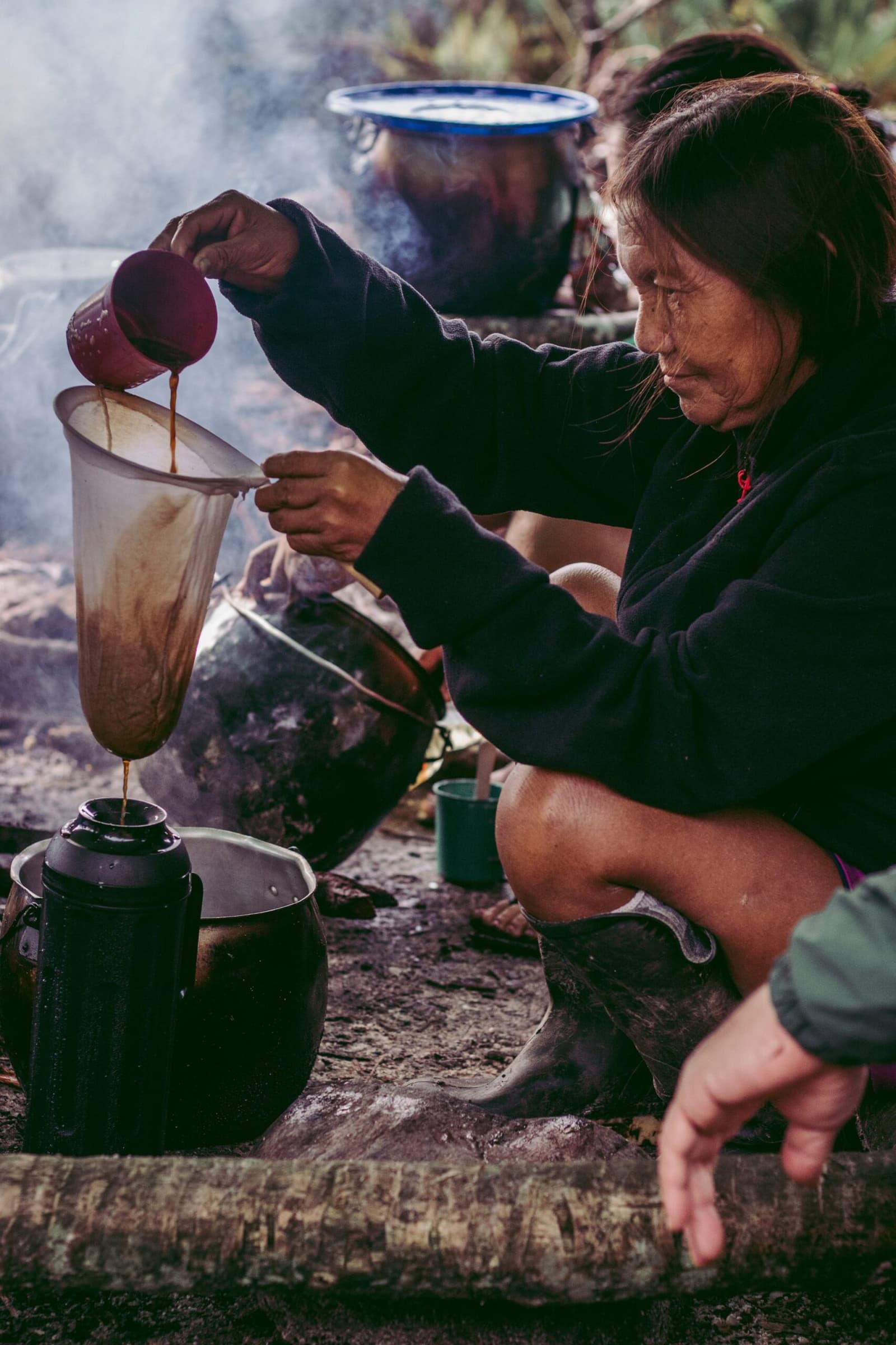 Koffie met de hand gefilterd, vuur achter haar, rook overal. Sommige gewoonten zijn gewoon goed. Een Yanomami-vrouw hurkt bij een open vuur en giet donkere vloeistof door een stoffen filter in een zwartgeblakerde ketel, omgeven door rook