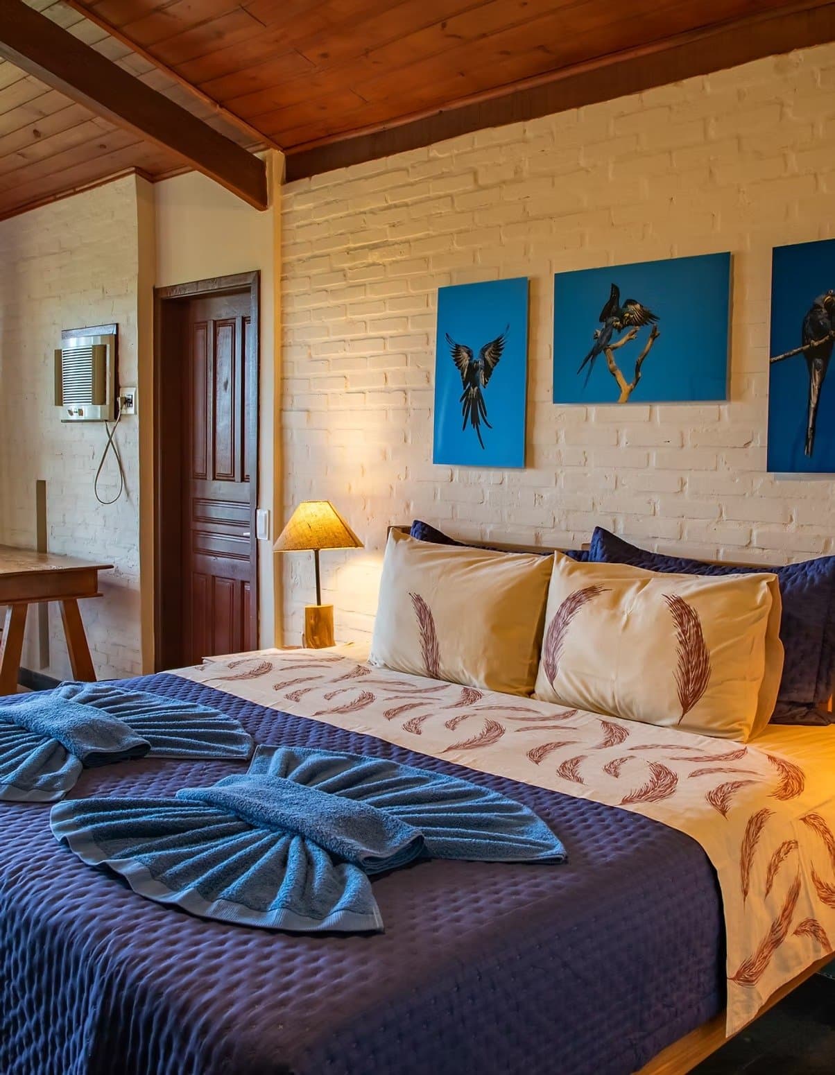 Des aras au mur, des plumes sur l'oreiller. Le lodge sait où il est. Chambre de lodge de safari dans le sud du Pantanal avec un grand lit habillé de linge imprimé de plumes, un couvre-lit matelassé bleu, des serviettes pliées en éventail, trois toiles bleues représentant des aras, une lampe de chevet en bois et un mur de briques blanches sous un plafond à poutres apparentes