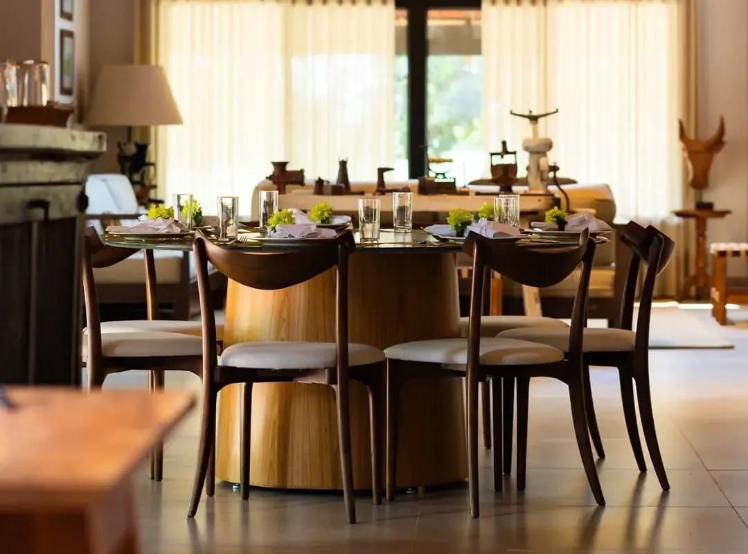 Un dîner bien mérité après une journée dans les zones humides, servi avec soin. Table à manger ronde élégamment dressée avec des serviettes en lin pliées, des verres et de petits arrangements floraux verts dans l'intérieur chaleureux d'un lodge de safari au sud du Pantanal, avec des meubles en bois vintage et des rideaux transparents