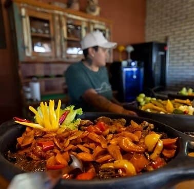 La cuisine pantaneira ne se presse pas, et le repas s'en ressent. Une poêle en fonte remplie de viande mijotée, de poivrons et d'oignons, garnie de lanières de poivron jaune, servie dans un lodge de safari dans le sud du Pantanal pendant qu'un cuisinier s'affaire en arrière-plan