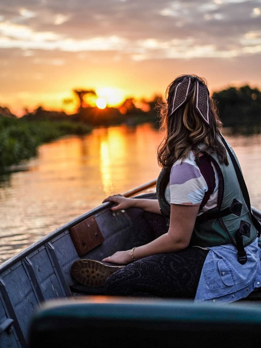 L'heure dorée, un canoë, et plus personne pour se dépêcher. Une femme portant un gilet de sauvetage est assise dans un petit canoë en bois, pagaie en main, regardant le soleil se coucher sur un canal tranquille bordé de végétation basse lors d'un safari dans le sud du Pantanal