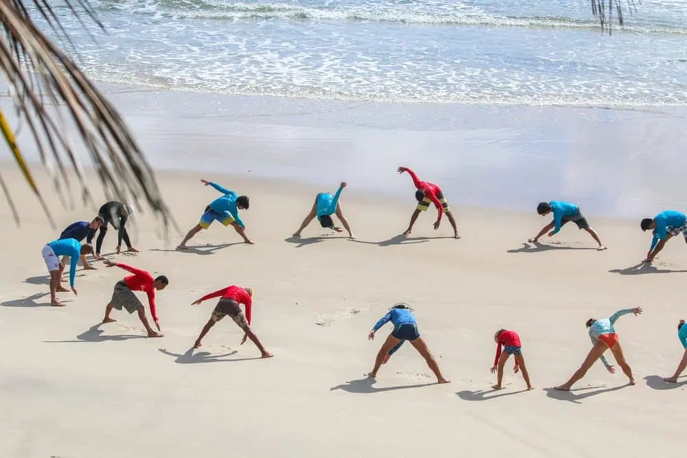 Eerst goed opwarmen, dan het water in. Een groep studenten in rash guards doet rekoefeningen op een wit zandstrand voor hun surflessen in Itacaré, met rustige oceaangolven op de achtergrond.