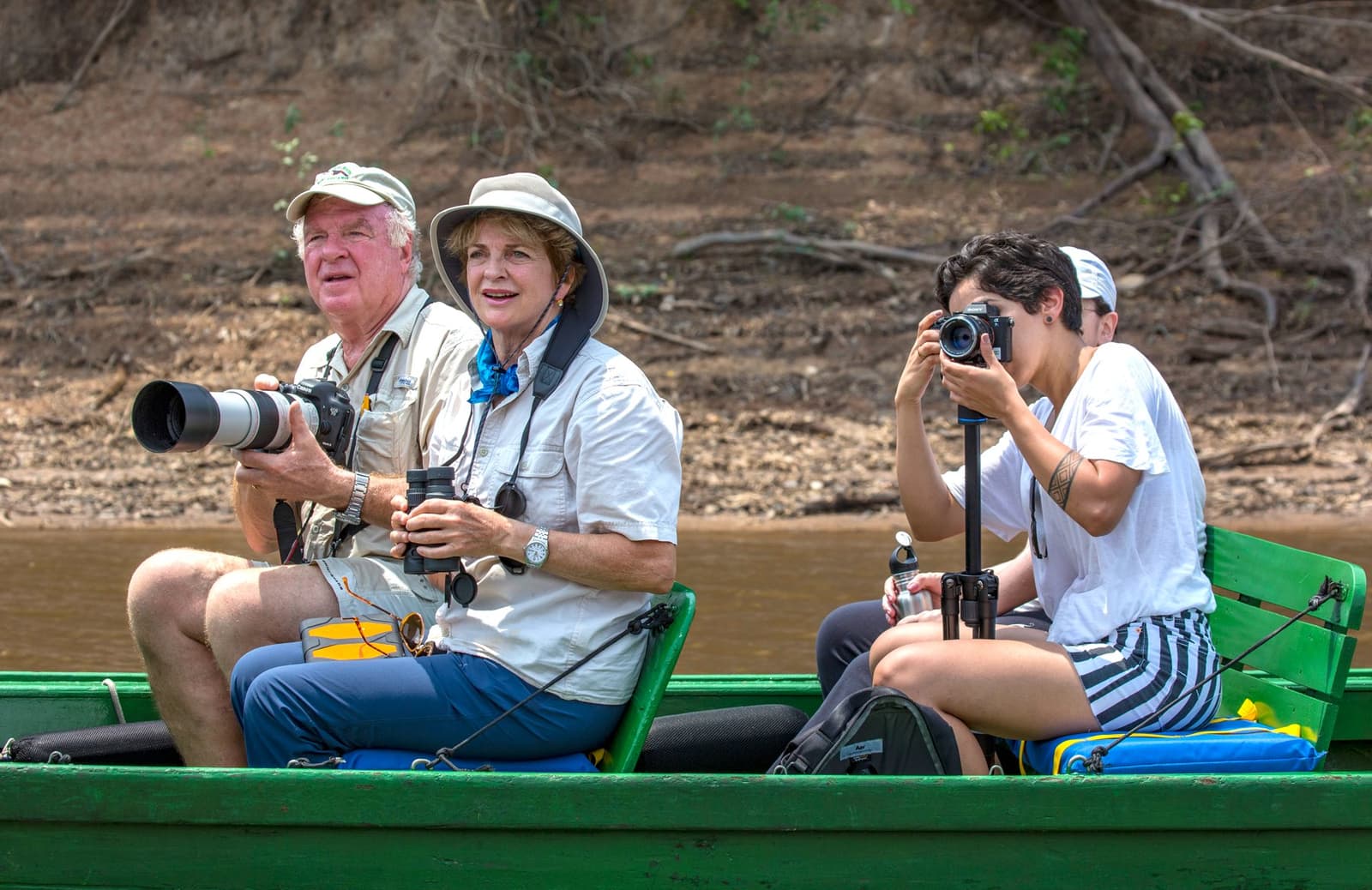 Lenzen gericht, rivier voor je, en de volgende ontdekking wacht om de bocht. Photo: Daniel Forster Drie reizigers op een groene houten boot langs een rivieroever tijdens een Tucano Amazon-riviercruise: een man met een teleobjectief, een vrouw met een verrekijker en een jongere persoon die fotografeert met een camera op een monopod