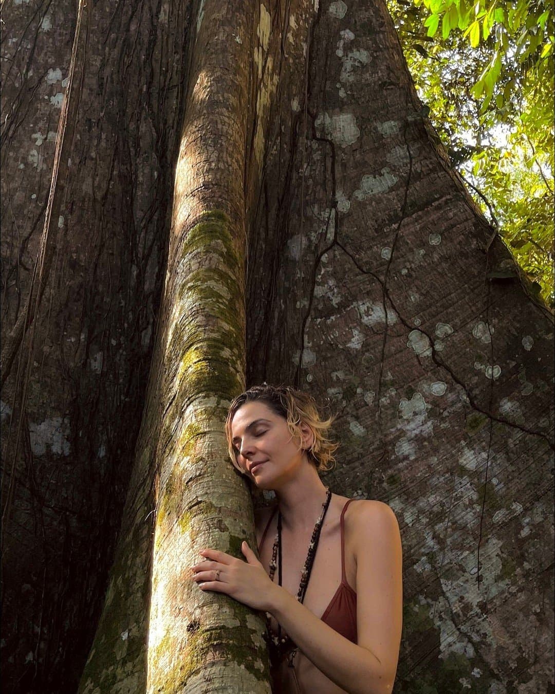 Some trees earn a hug. This one has been waiting centuries. A woman with eyes closed peacefully rests her cheek against the moss-covered trunk of a towering ancient tree during an Amazon River Cruise Brazil experience, its massive buttress roots spreading behind her