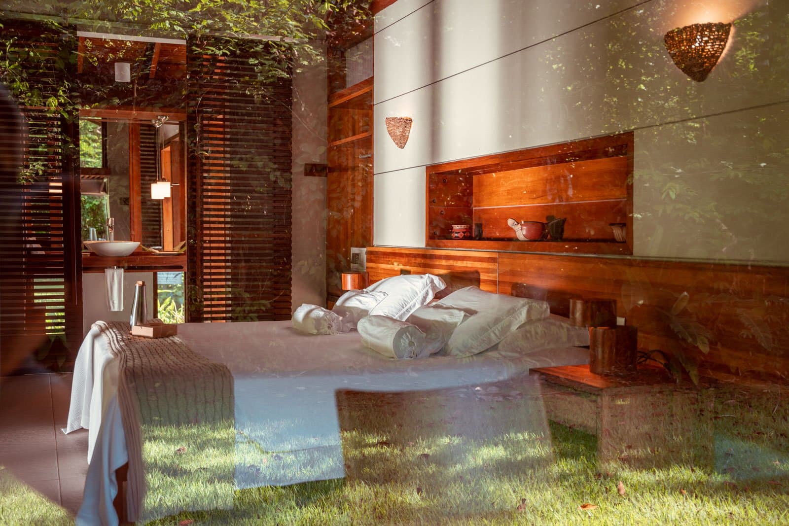 The forest reflects in the glass, just to remind you it never really goes away. Photo: Samuel Melim Interior of a jungle lodge bungalow viewed through a glass wall, showing a double bed with white pillows and a knitted throw, warm timber headboard with built-in shelving, woven wall sconces, and a vanity area with wooden louvred shutters opening to lush green forest — comfortable accommodation for birdwatching in the Amazon rainforest