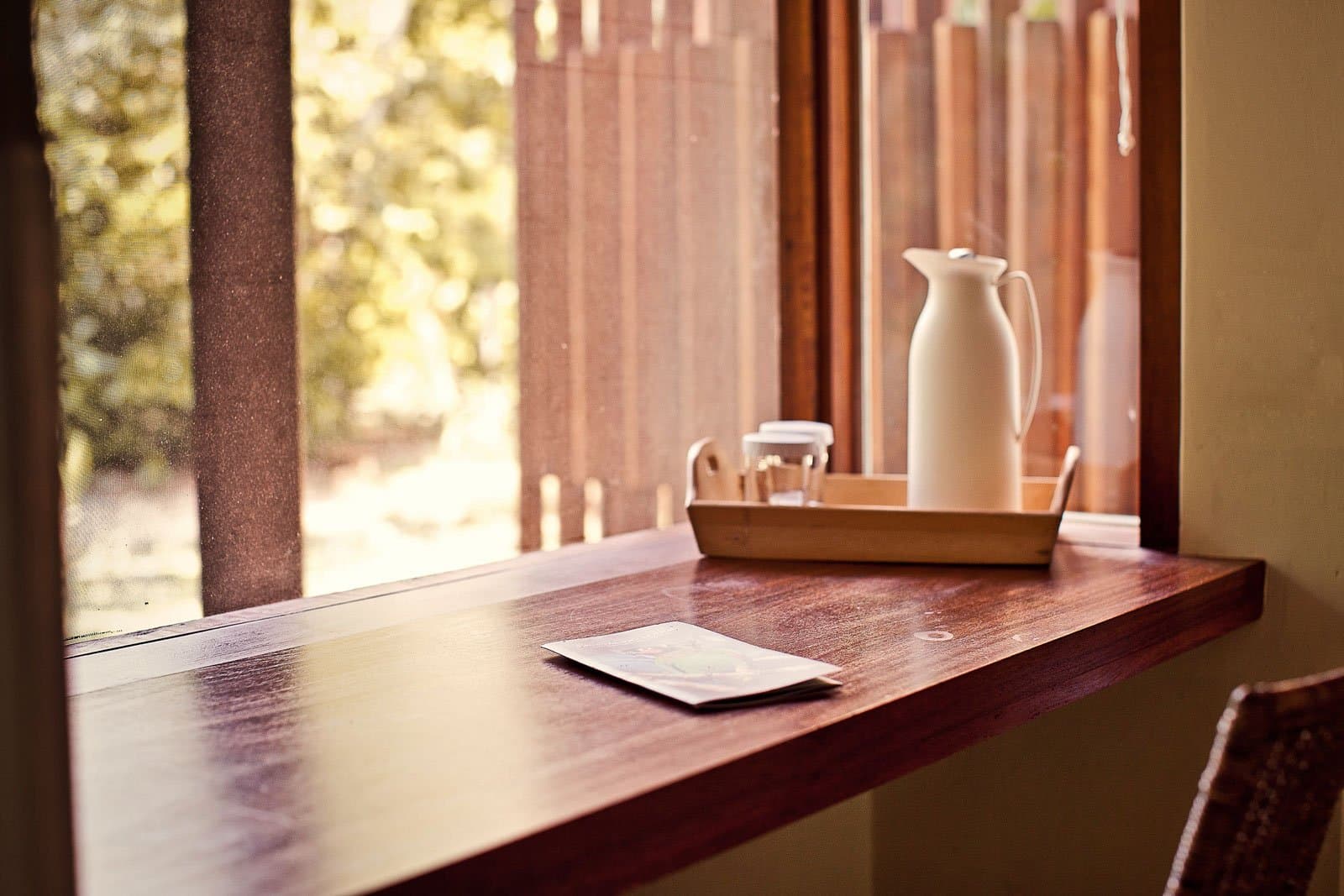 Quiet corner, hot coffee, trees just outside. The forest can wait a minute. Photo: Samuel Melim A white thermal carafe and glass on a wooden tray sit on a warm timber desk beside a screened window overlooking green forest, with a small card resting on the desk surface — a quiet corner inside a birdwatching in the Amazon rainforest lodge