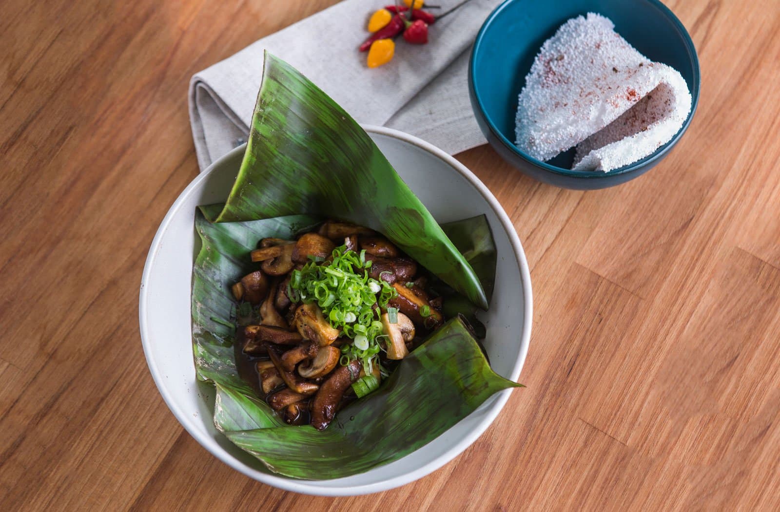 Champignons sauvages, feuille de bananier, farine de manioc. La forêt se met à table. Photo: Rjcastilho Champignons sautés garnis d'oignons verts servis dans une feuille de bananier dans un bol blanc, avec un bol bleu de farine de manioc saupoudrée de piment et de petits piments frais sur une table en bois — repas dans un lodge d'observation des oiseaux en Amazonie