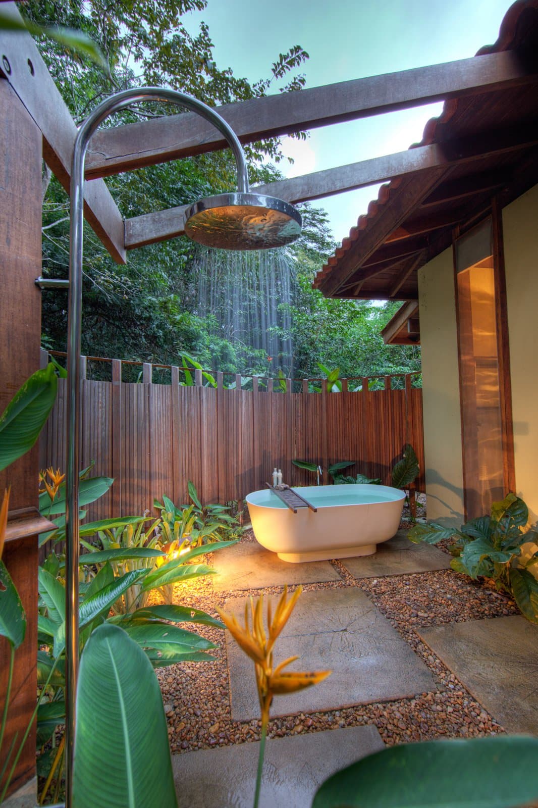 Hot soak, open sky, and the forest a few feet away. Not bad after a morning on the trails. Photo: Samuel Melim A freestanding soaking tub filled with water sits in an open-air garden courtyard surrounded by tropical plants and warm lighting, with a large rainfall showerhead in the foreground and dense forest canopy visible beyond the wooden fence — part of a birdwatching in the Amazon rainforest lodge experience