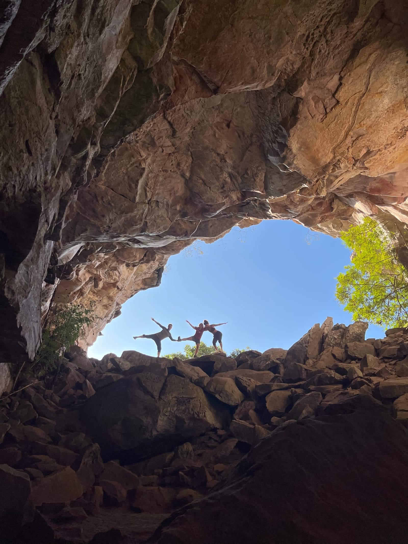 Eine Höhle entdecken und sofort in die Kriegerpose gehen, warum auch nicht. Zwei Trekker in Yoga-Posen als Silhouetten am Eingang einer großen Sandsteinhöhle auf dem Chapada-Diamantina-Trek, blauer Himmel und grüne Baumkronen durch den Felsbogen sichtbar