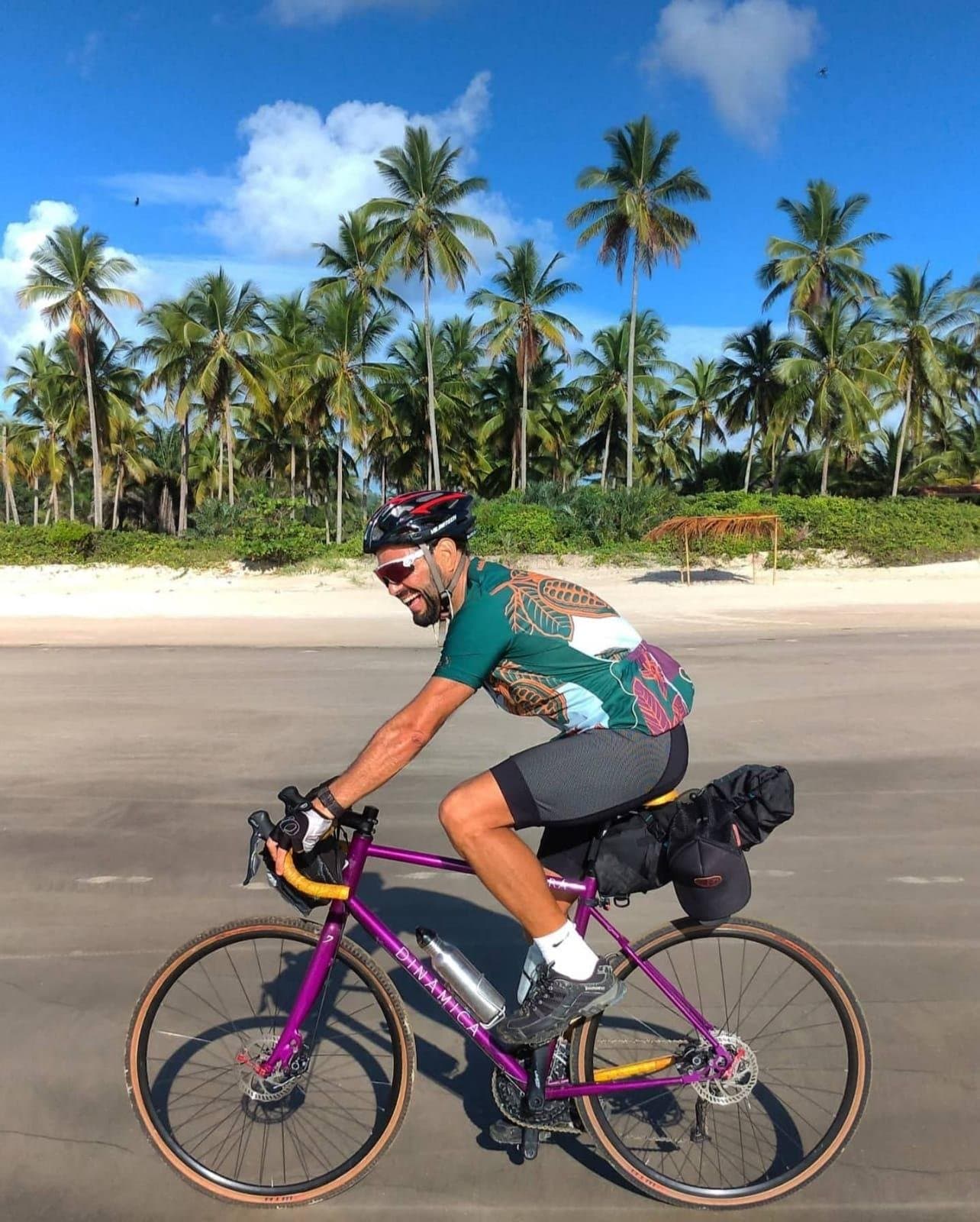 Aquele sorriso é de quem sabe que a estrada ainda tem muito para dar. Um ciclista sorridente em uma gravel roxa carregada pedala por uma estrada litorânea ladeada de coqueiros e praia de areia branca sob um céu azul intenso, retratando a essência do cicloturismo na Bahia.