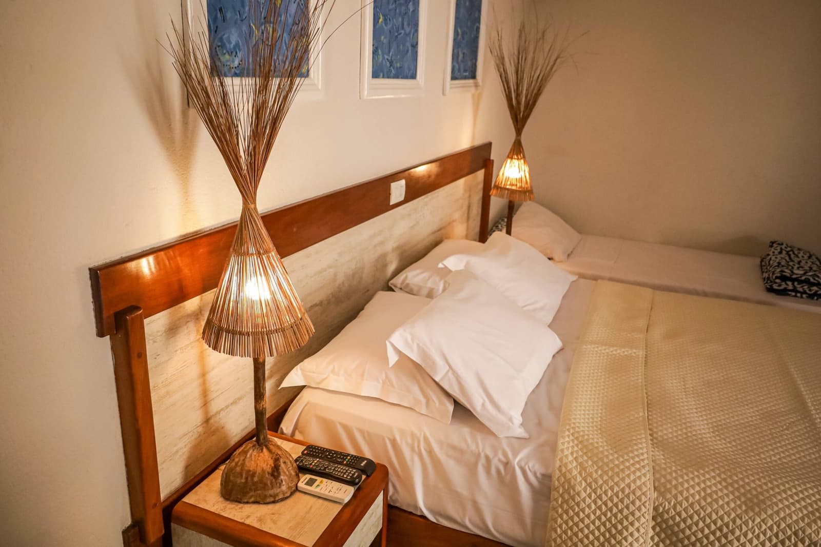 Quarto simples, sono de pedra, e amanhã a estrada chama de novo. Um quarto aconchegante e simplesmente mobiliado de uma pousada na rota de ciclismo Bahia Cocoa Route, com duas camas vestidas de lençóis brancos, abajures artesanais de ratan sobre criado-mudo de madeira e janelas decorativas azuis acima da cabeceira