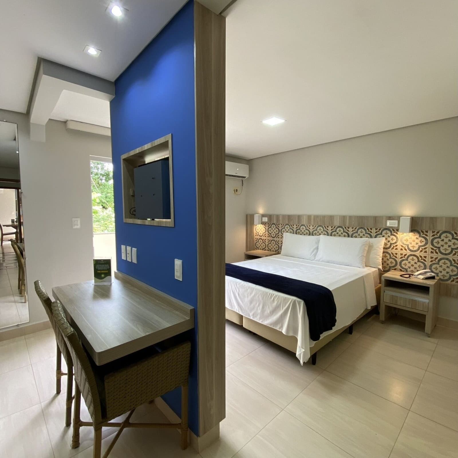 Conforto simples e bem resolvido, do jeito que a noite pede. Quarto de hotel limpo com cama de casal, cabeceira com azulejos estampados, escrivaninha de madeira, cadeiras de vime e parede de destaque azul com televisão embutida