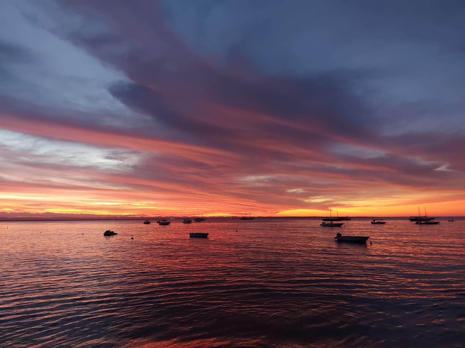 O fim de tarde na baía de Itacaré não pede nenhuma legenda. Pôr do sol em tons de vermelho e laranja intensos sobre uma baía calma com silhuetas de barcos de pesca ancorados, visto da orla de Itacaré na rota ciclística do cacau na Bahia