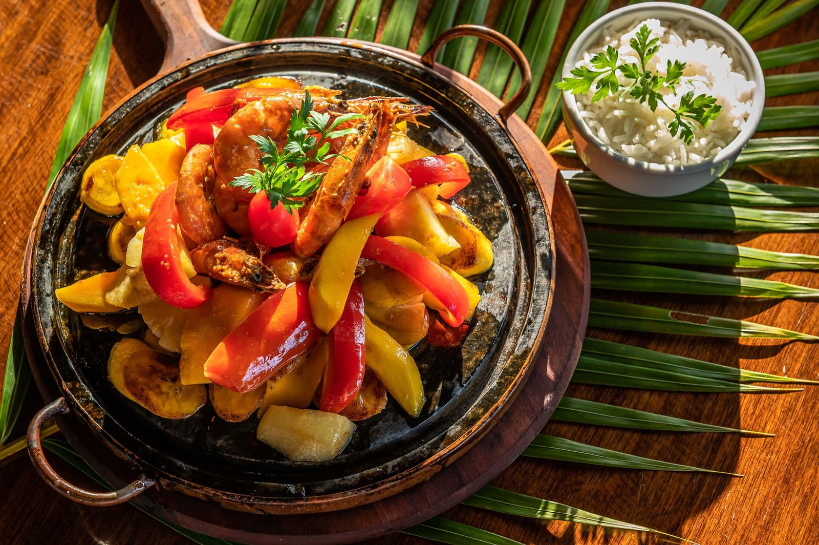 Pedalar pela Bahia abre um apetite honesto. Essa frigideira entende o recado. Frigideira de barro fumegante com camarões inteiros grelhados, pimentões vermelhos e amarelos e um potinho de arroz branco com salsinha, servida sobre folhas de palmeira em tábua de madeira