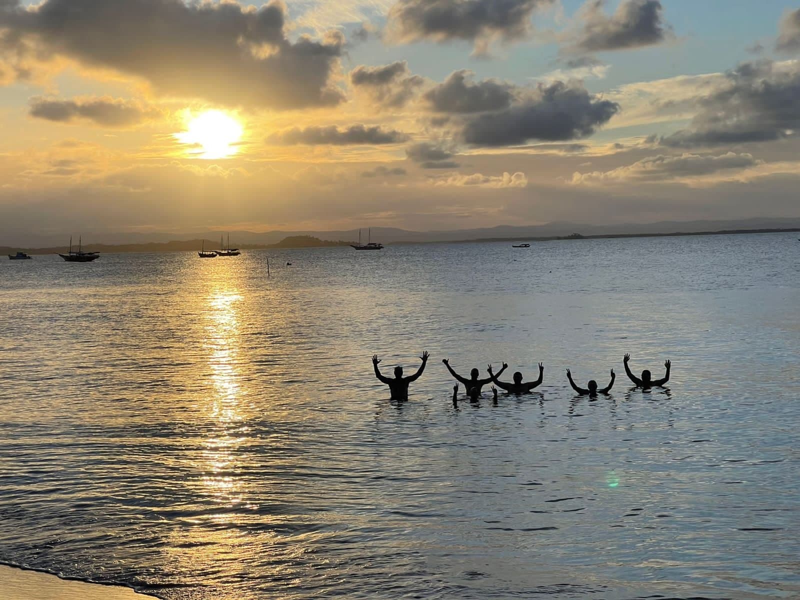 Braços pro alto, sol se pondo, o Rio das Contas sabe fechar o dia. Cinco silhuetas na água com os braços erguidos celebrando um pôr do sol dourado sobre uma baía calma com barcos ancorados ao fundo
