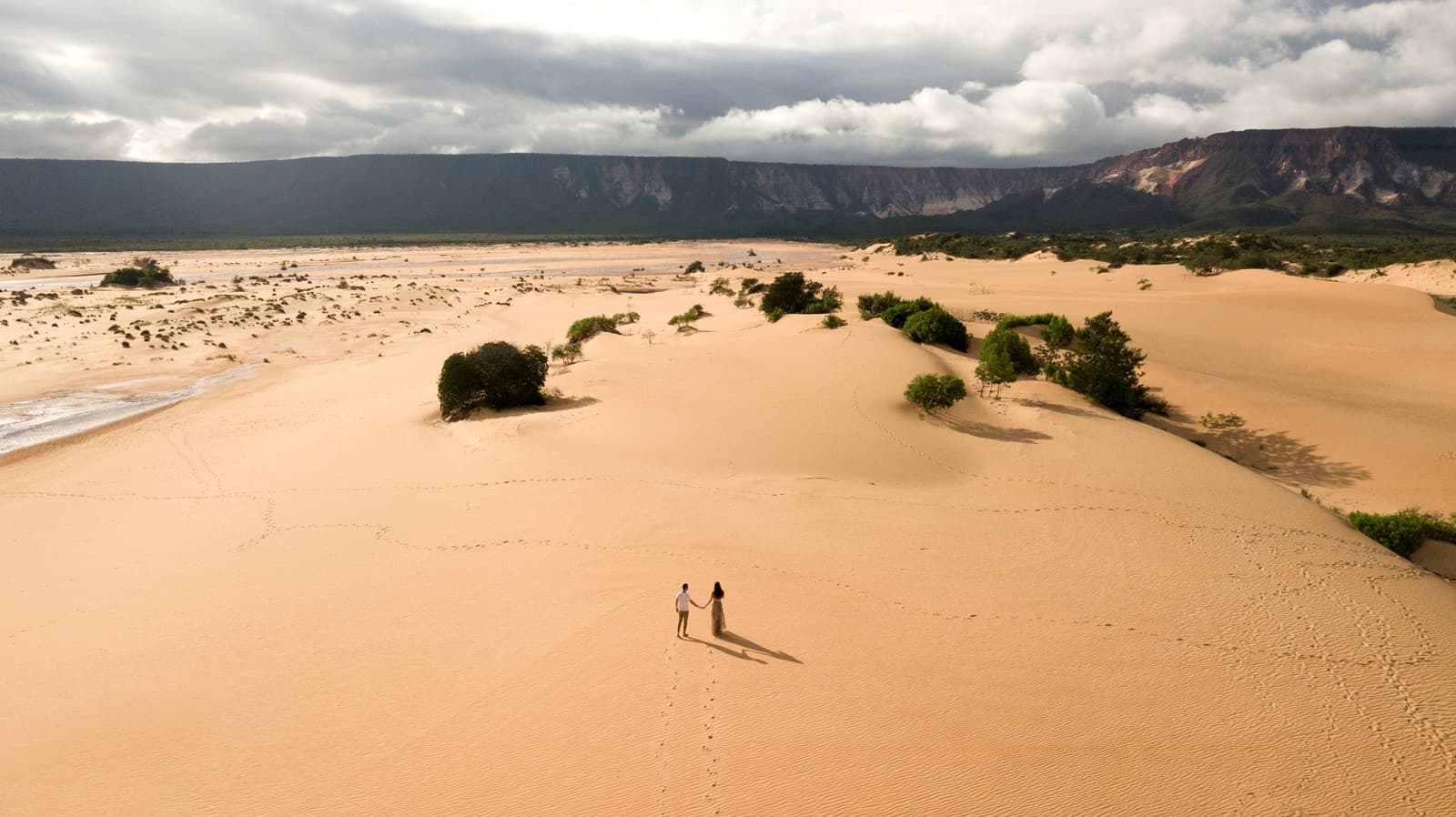 Dois, as dunas imensas, e pegadas que contam a história sem precisar de legenda. Photo: FG Trade Vista aérea de um casal de mãos dadas sobre as vastas dunas douradas do Jalapão, com arbustos verdes, a margem de um rio e uma escarpa de chapada sob um céu carregado — um momento íntimo em plena aventura no Jalapão.