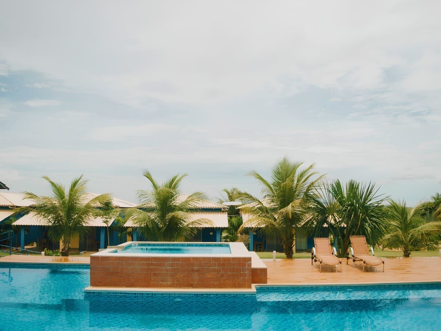 Depois de trilha e fervedouro, essa piscina merece cada metro percorrido. Piscina externa com hidromassagem elevada e duas espreguiçadeiras ao lado de palmeiras em uma pousada no Jalapão, com bangalôs azuis ao fundo sob céu parcialmente nublado