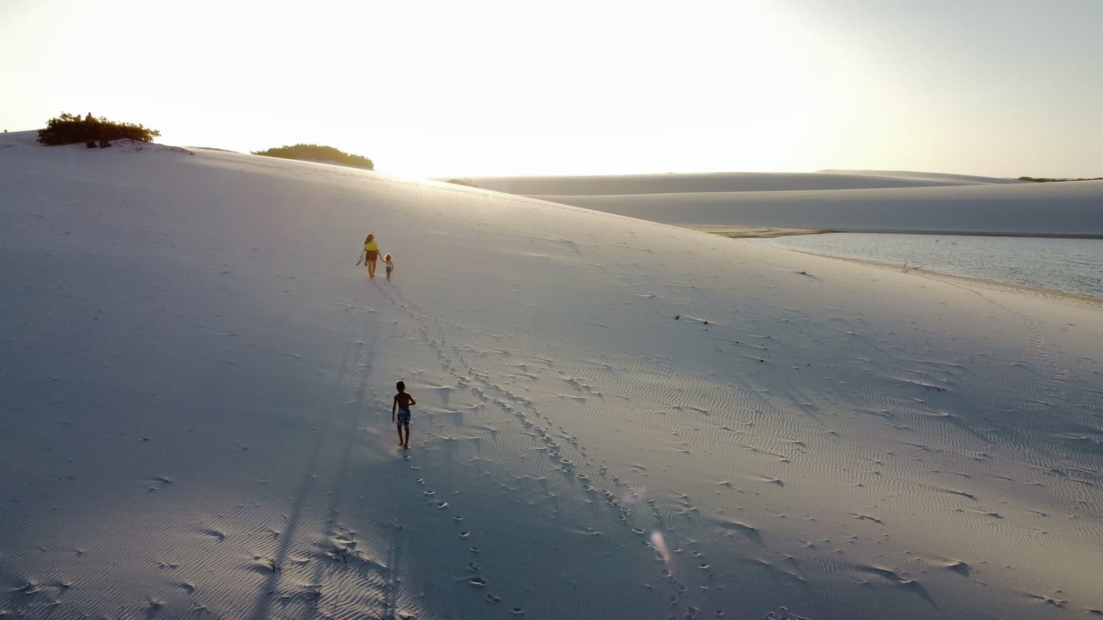 A duna mede a vontade, não a altura de quem sobe. Um adulto e duas crianças sobem uma enorme duna de areia branca ao lado de uma lagoa calma durante o horário dourado nos Lençóis Maranhenses