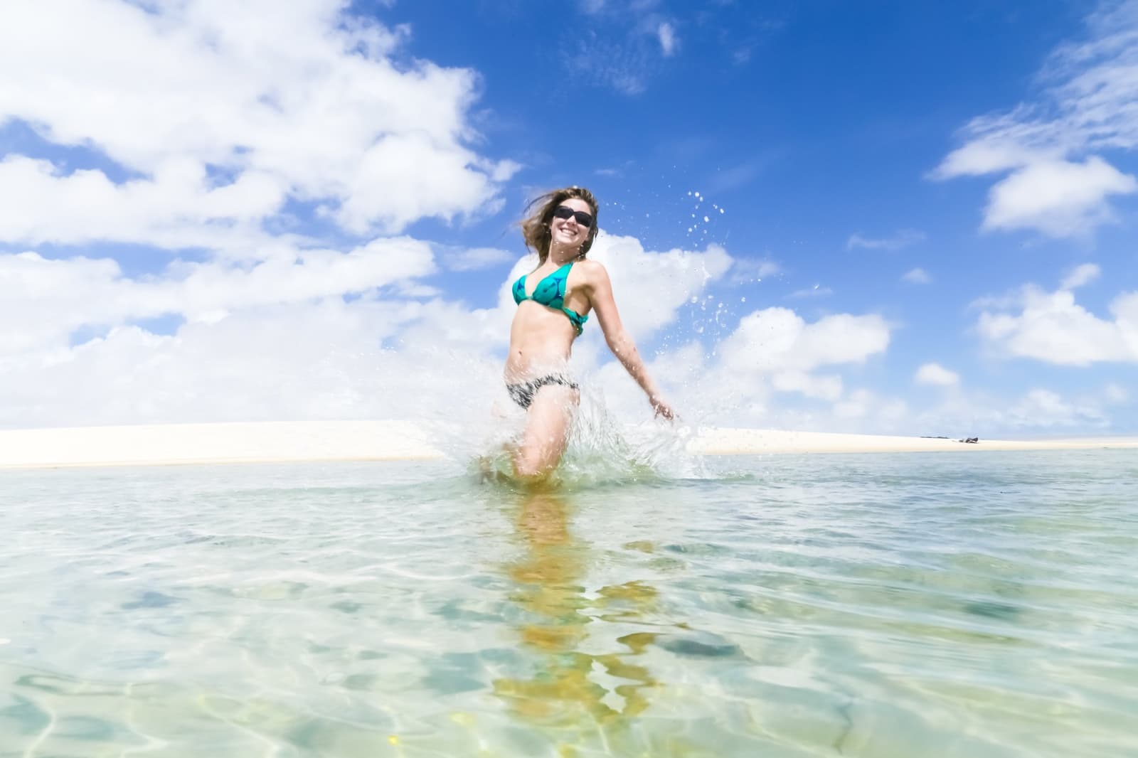 Água fria, areia quente, e a vida fazendo sentido. Uma mulher sorridente de biquíni verde-azulado espirra água cristalina rasa de lagoa com dunas de areia branca e céu azul intenso ao fundo nos Lençóis Maranhenses