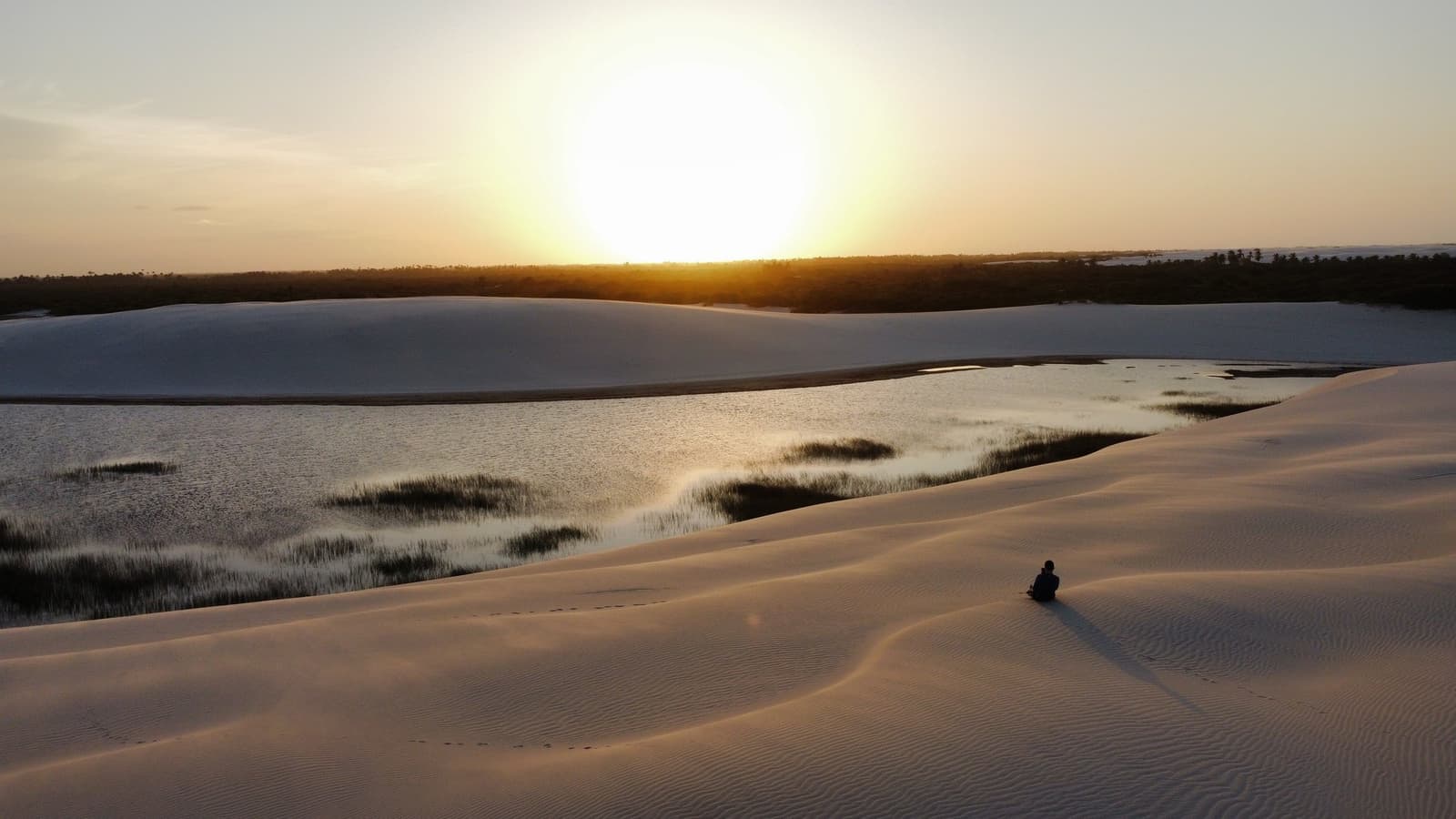 A melhor poltrona dos Lençóis não tem encosto, só vista. Uma figura solitária sentada sobre dunas de areia branca ondulada contempla uma lagoa rasa com tufos de vegetação durante um pôr do sol dourado, cena marcante do roteiro completo dos Lençóis Maranhenses