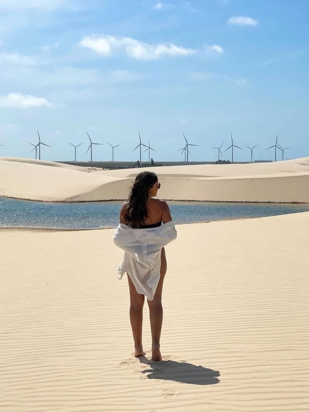 Areia, água e vento, os Lençóis Maranhenses têm um jeito todo próprio de encantar. Uma mulher de camisa branca está descalça sobre dunas de areia branca ondulada, olhando para uma lagoa azul brilhante, com uma fileira de turbinas eólicas se estendendo pelo horizonte — uma cena marcante do roteiro completo pelos Lençóis Maranhenses