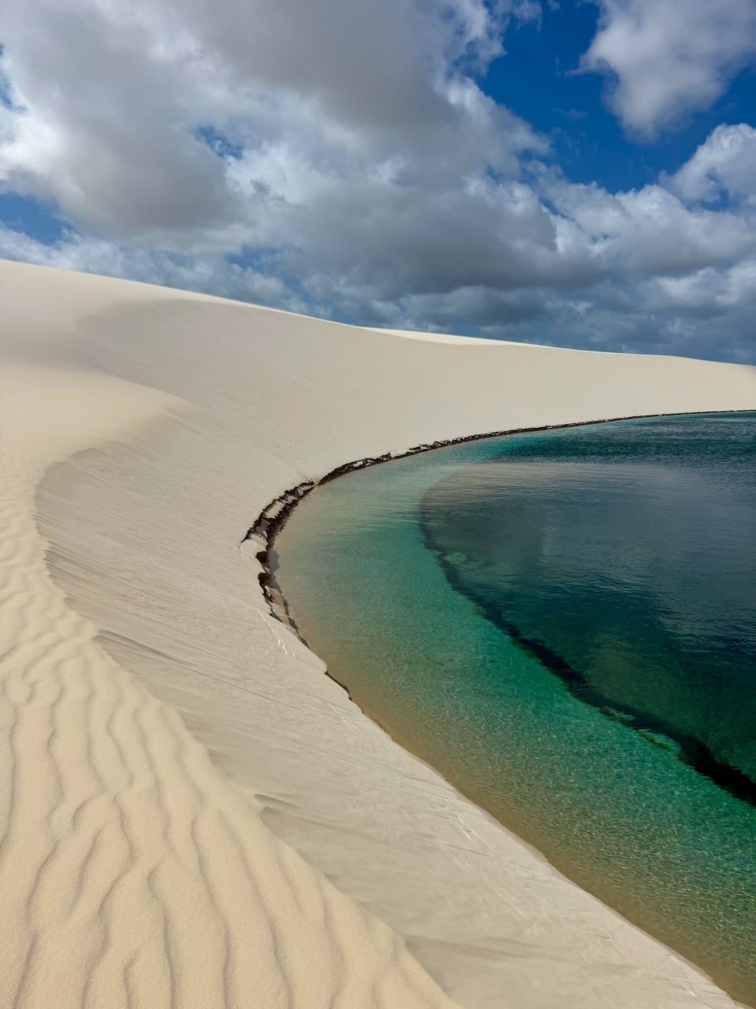 Du sable aussi blanc, de l'eau aussi claire, les Lençóis Maranhenses tiennent leurs promesses. Un lagon turquoise en forme de croissant niché contre une grande dune de sable blanc sous un ciel bleu partiellement nuageux à Lençóis Maranhenses, avec un rivage peu profond passant du bleu limpide au vert profond