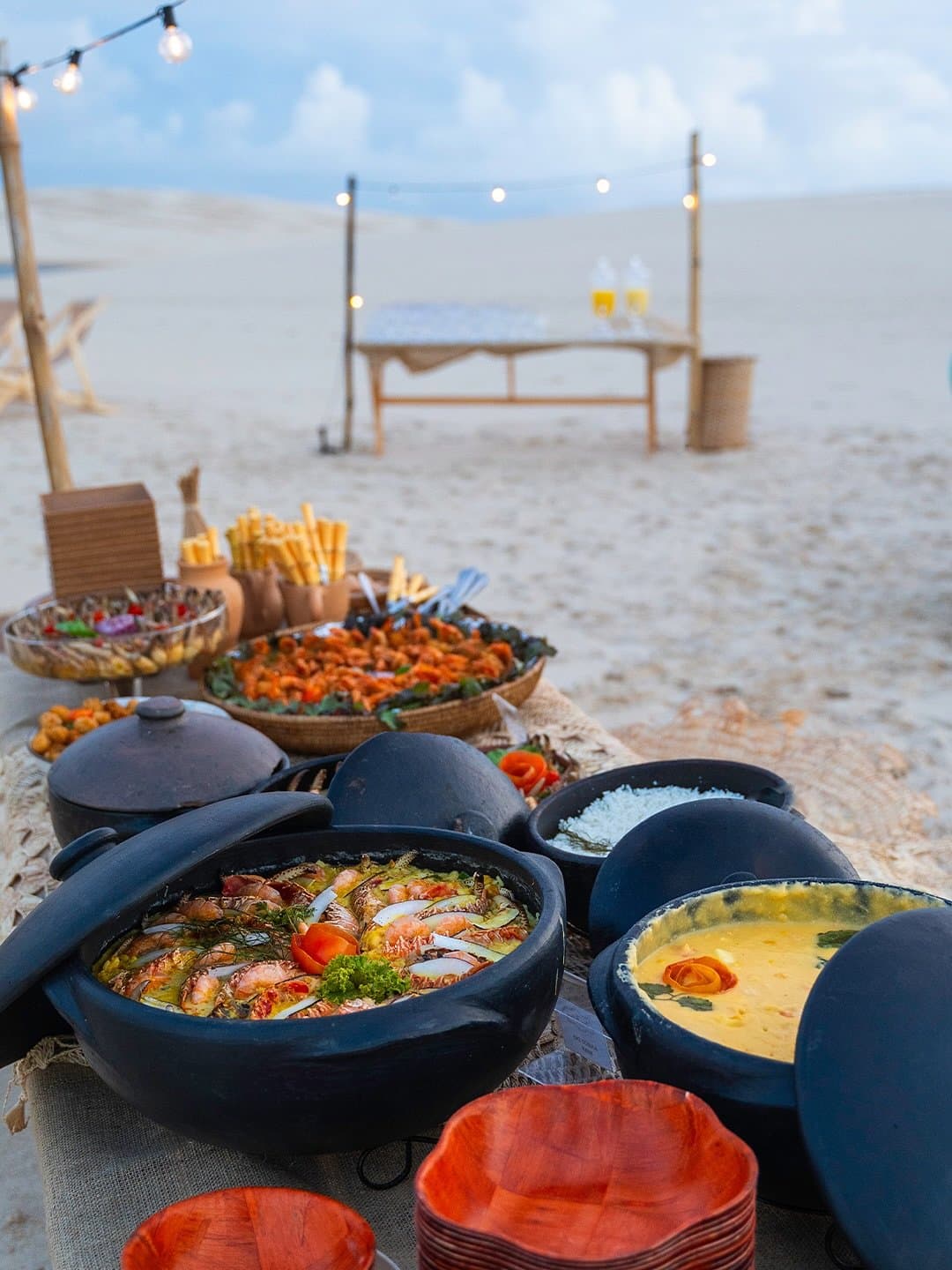 Un festin sur le sable, là où la salle à manger n'a pas de murs. Un festin de plats de fruits de mer dans des casseroles en argile traditionnelles, dont une moqueca aux crevettes et une sauce crème dorée, disposés sur une table rustique dans le sable blanc des dunes sous des guirlandes lumineuses au crépuscule