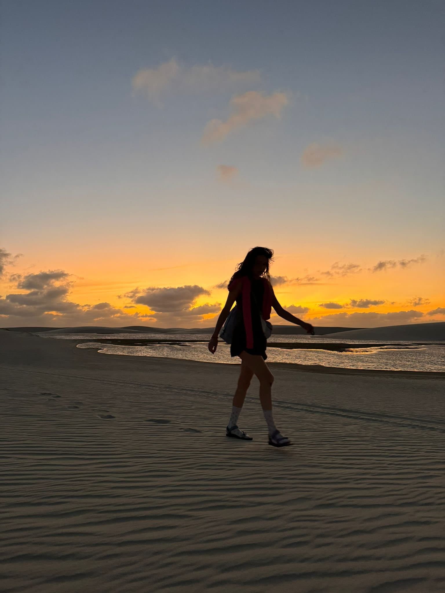 Le sable garde vos empreintes juste assez longtemps pour que ça compte. Silhouette d'une femme marchant sur des dunes de sable ondulées au coucher du soleil à Lençóis Maranhenses Santo Amaro, avec une lagune peu profonde et un ciel orangé en arrière-plan
