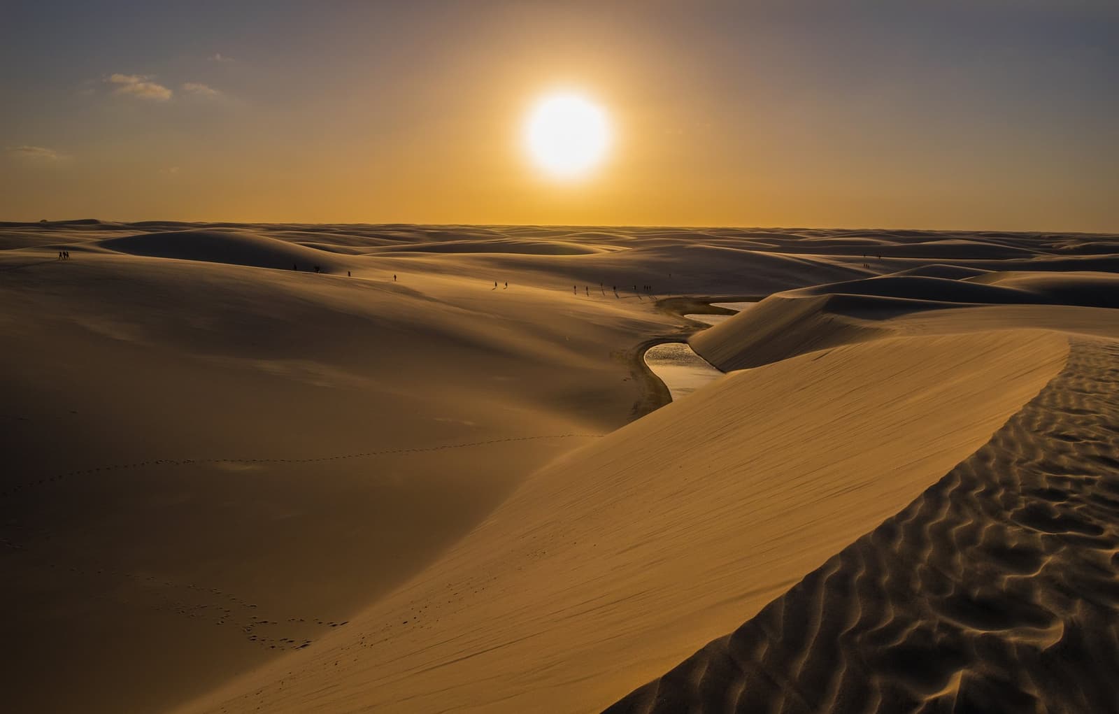 O sol se despede do dia do jeito que só os Lençóis sabem receber. Extensas dunas de areia branca do trek pelos Lençóis Maranhenses banhadas pela luz âmbar do sol baixo, com uma pequena lagoa de água doce serpenteando entre as dunas e silhuetas minúsculas de caminhantes espalhadas pelo cume ao fundo