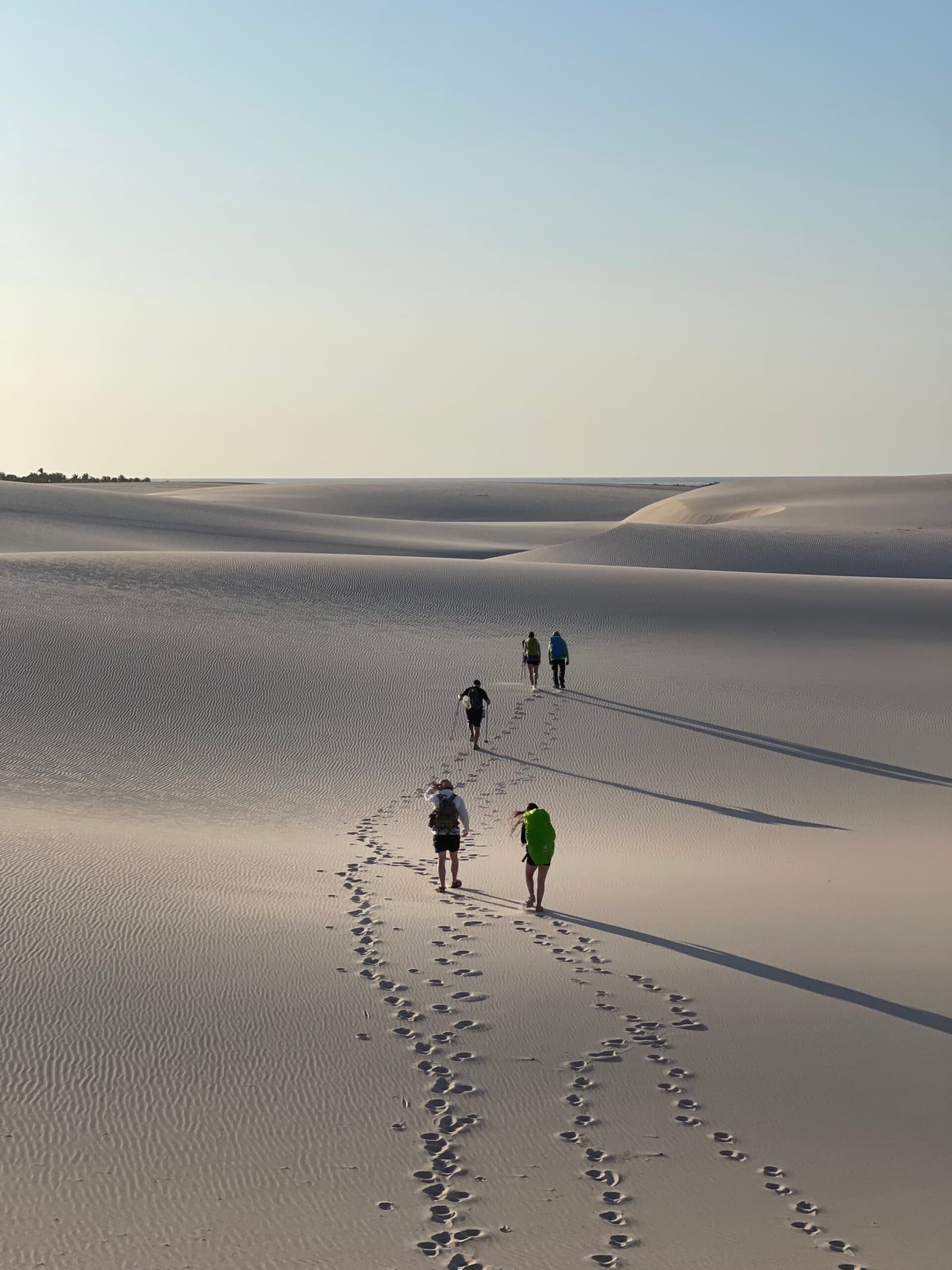 Quatro pessoas, um mar de areia branca, e a trilha toda para chamar de sua. Cinco caminhantes com mochilas atravessam as dunas brancas e onduladas dos Lençóis Maranhenses no trek pelo parque, deixando pegadas na areia sob um céu claro da manhã