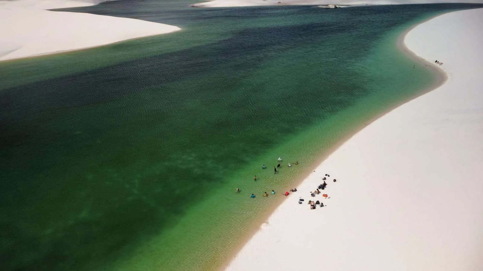 Figures minúsculas, lagoa enorme, aqui a escala resolve qualquer discussão. Vista aérea de uma grande lagoa de água doce no trek pelos Lençóis Maranhenses, com águas verde-escuras e turquesa contornando dunas de areia branca, e um pequeno grupo de visitantes entrando pela beira