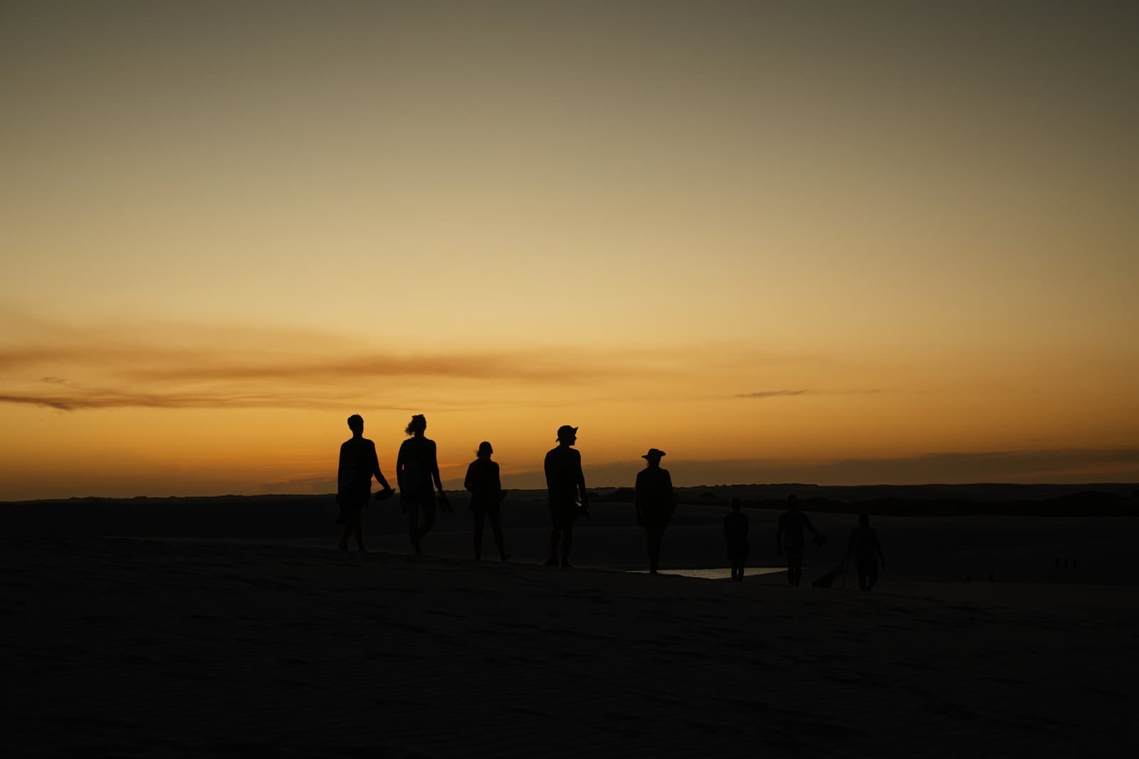 Le sable garde la chaleur, le ciel garde les couleurs. Silhouettes d'un groupe de randonneurs marchant sur les dunes ouvertes des Lençóis Maranhenses au coucher du soleil doré