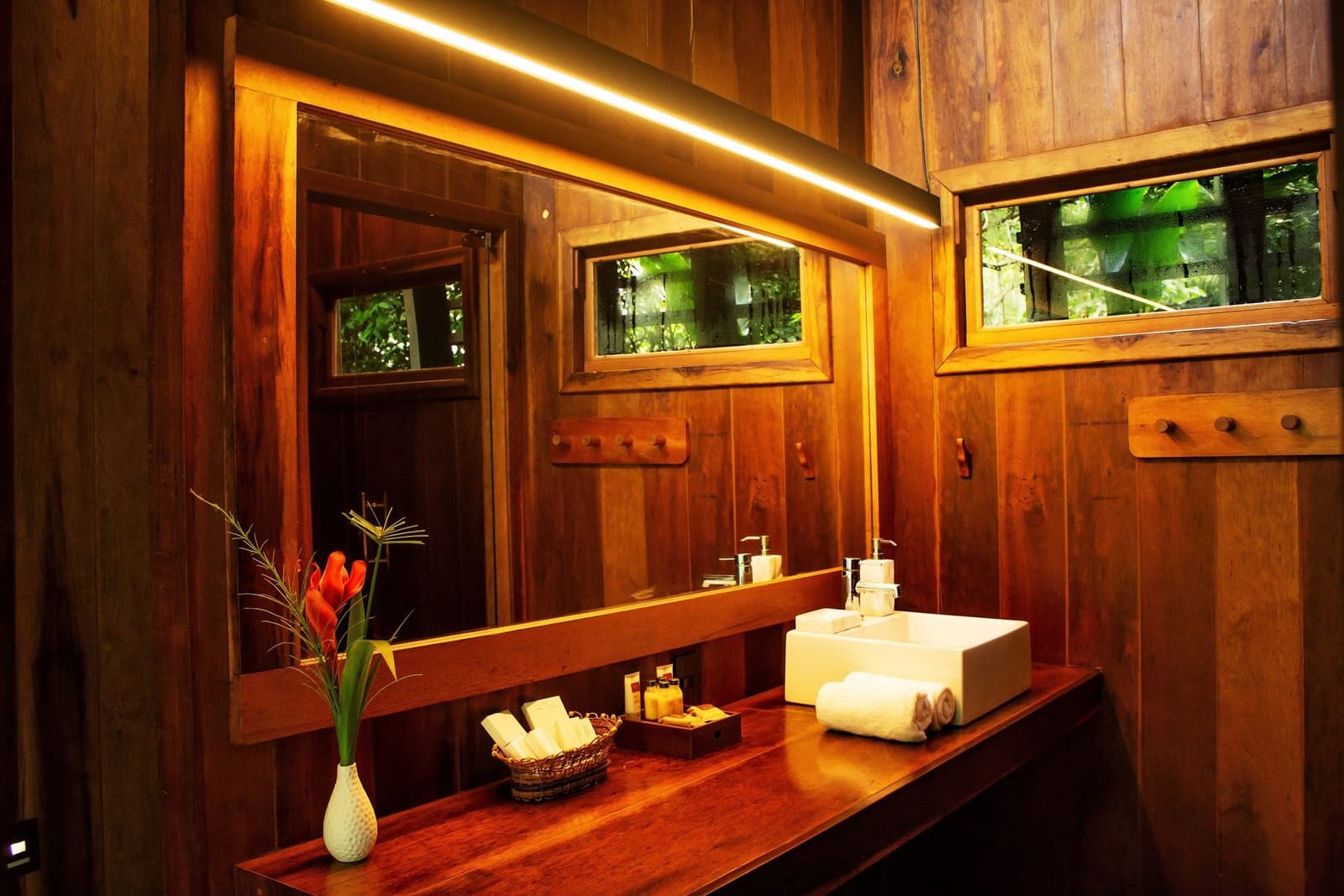 Le bois chaud, une fleur rouge, et la forêt qui regarde par la fenêtre. Salle de bain chaleureuse au Mirante do Gavião avec un plan de travail en bois poli, une vasque blanche, des fleurs tropicales rouges et des fenêtres donnant sur la jungle