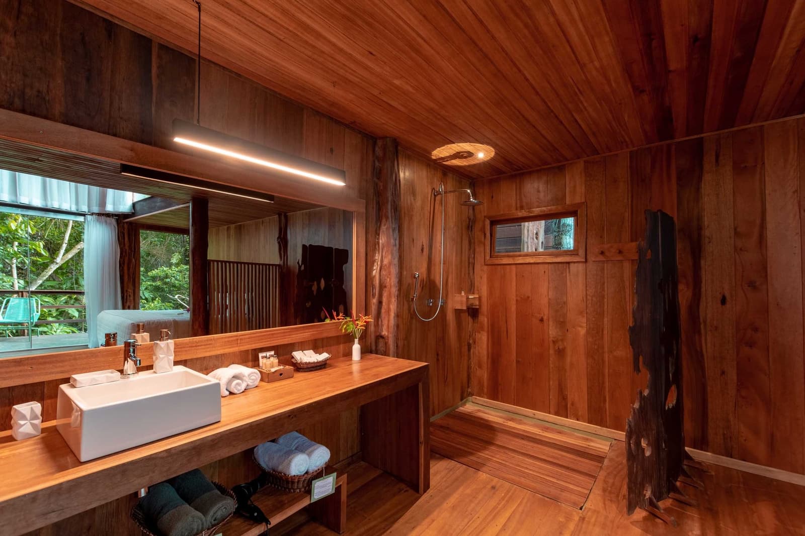 Du bois, de la lumière douce, et la forêt à portée de regard. Salle de bain entièrement en bois au Mirante do Gavião avec vasque posée, douche à effet pluie, serviettes pliées et vue sur la jungle par la fenêtre