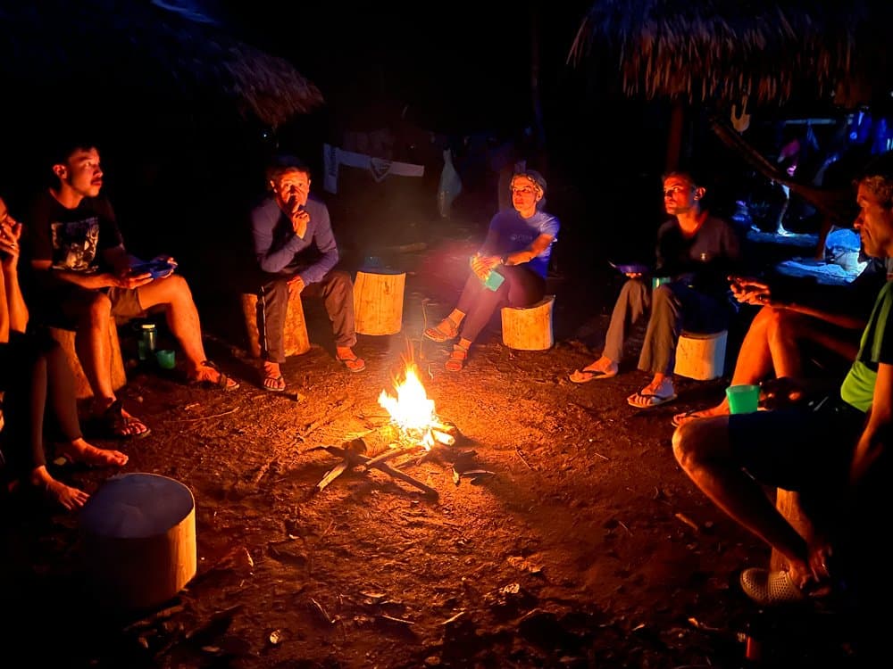 Het vuur zegt niets, maar iedereen zit er toch naar te luisteren. Een groep reizigers op de Pico da Neblina Expedition zit 's nachts op boomstronken in een kring rond een klein kampvuur, onder een rieten afdak in het Amazone-regenwoud